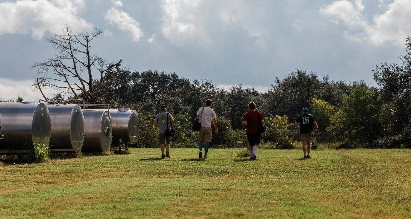 Four disc golfers walking across the open grass at Live Oak Brewing, past the brewery's steel tanks